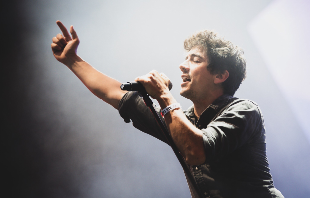 Ein Mann in schwarzem Hemd und Jeans singt leidenschaftlich in ein Mikrofon auf einer hell erleuchteten Bühne, mit einem engagierten Publikum, das im Hintergrund klatscht und jubelt.