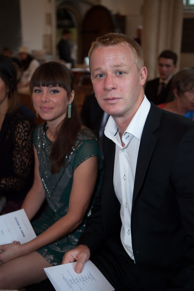 Ein Mann in schwarzem Mantel und weißem Hemd steht rechts und hält Karten, neben einem Mädchen in grünem Kleid, das ebenfalls Karten hält.