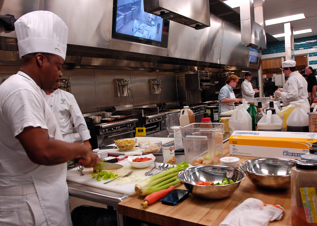Eine Gruppe von Köchen in einer Küche, mit einem im Vordergrund in einer weißen Schürze und Mütze, der ein Messer hält, umgeben von Utensilien, Schüsseln, Gemüse, einem Handy, Flaschen und anderen Gegenständen, mit einem Fernseher und Deckenleuchten im Hintergrund.