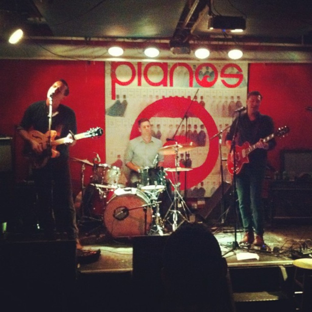 Drei Männer auf der Bühne: einer spielt Schlagzeug in der Mitte, zwei spielen Gitarre und singen links und rechts, mit einem Banner an der Wand und Lampen an der Decke.