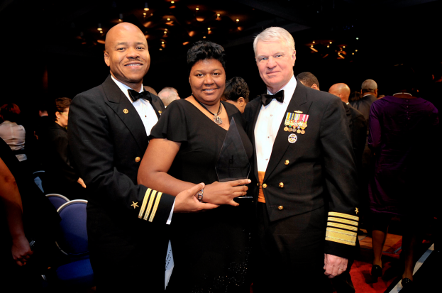 Zwei Männer in schwarzen Blazern und weißen Hemden stehen neben einer Frau in einem schwarzen Kleid, die eine Trophäe hält, auf einem roten Teppich, mit Menschen und beleuchteter Decke im Hintergrund.