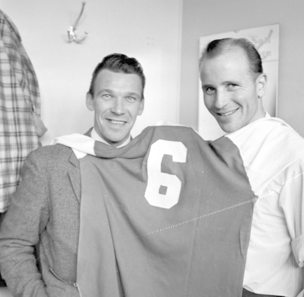 Zwei Männer in Schwarz-Weiß, die strahlend ein Fußballtrikot hochhalten, mit einer Tafel und einem Bügel an der Wand dahinter.