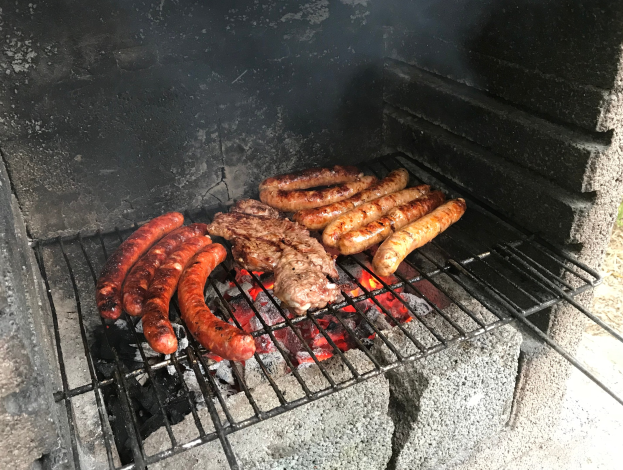 Ein Grill mit Würstchen und Fleisch darauf, umgeben von brennender Kohle und Feuer, umgeben von Gras im Freien.