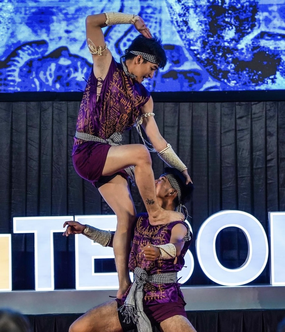 Zwei Performer in purpurfarbenen Kleidern führen einen akrobatischen Hebemanöver auf der Bühne vor einem Publikum aus, mit einem schwarzen Vorhang und einer Bildschirmhintergrund.