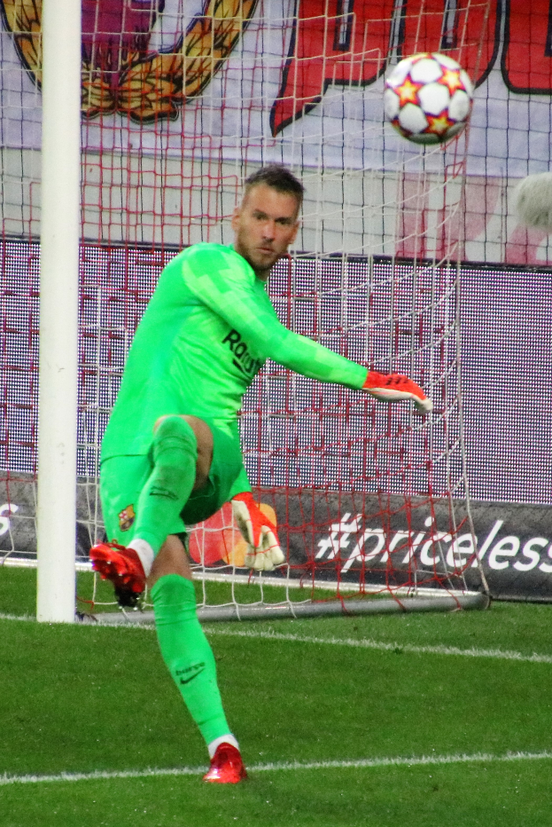 Ein Fußballspieler in grüner Uniform fängt einen Ball vor einem Tor mit Netz und einem Banner im Hintergrund, trägt Fußballhandschuhe.