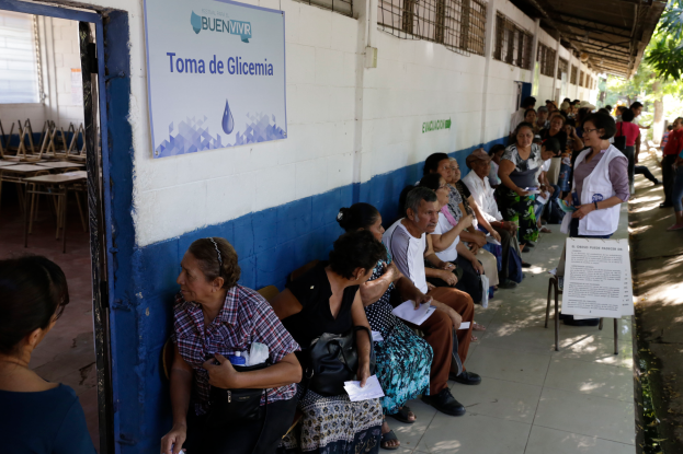 Eine Gruppe von Menschen sitzt auf Bänken vor einem Gebäude mit einer Texttafel an der Wand, einer Tür links und Bäumen im Hintergrund und wartet darauf, bei einer Wahllokal in Guadalajara, Mexiko, ihre Stimmen abzugeben.