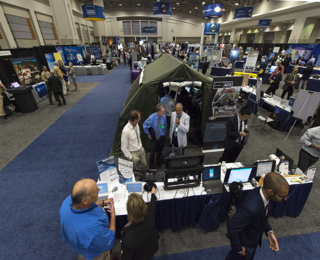 Eine große Gruppe von Menschen auf einer Karriere-Messe um einen Tisch mit Laptops, Monitoren und Papieren versammelt, mit einem Zelt, Plakaten, Deckenleuchten und Fenstern im Hintergrund.