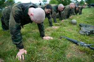 Eine Gruppe von Männern in Militäruniformen, die Liegestütze auf Gras machen, mit einer Waffe, einem Helm und anderen Gegenständen drumherum, mit Bäumen, Lichtern, Fahrzeugen und einem klaren blauen Himmel im Hintergrund.