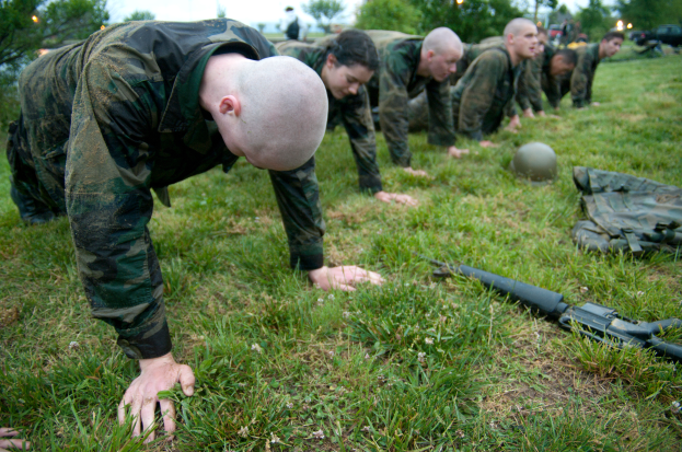 Eine Gruppe von Männern in Militäruniformen, die Liegestütze auf Gras machen, mit einer Waffe, einem Helm und anderen Gegenständen drumherum, mit Bäumen, Lichtern, Fahrzeugen und einem klaren blauen Himmel im Hintergrund.