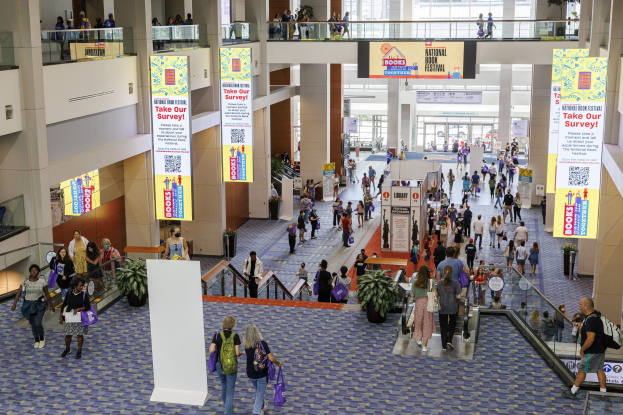 Eine große Gruppe von Menschen geht durch ein großes Gebäude mit Transparenten, Topfpflanzen, Rolltreppen, Geländern, Säulen, Texttafeln, Glastüren und anderen Gegenständen, einige tragen Taschen, wahrscheinlich beim Nationalen Buchfest.