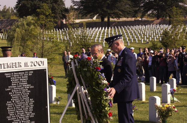 Eine Gruppe von Menschen versammelt sich um ein Grab auf einem Friedhof, während ein Mann in einer Militäruniform einen Kranz niederlegt, mit Blumensträußen, einem Textschild und umgebender Vegetation im Hintergrund.