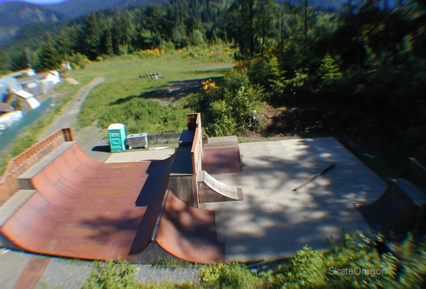 Eine Skaterbahn mit Kisten auf der linken Seite, Bäumen, Gras, Pflanzen und Blumen im Hintergrund und Text unten rechts.
