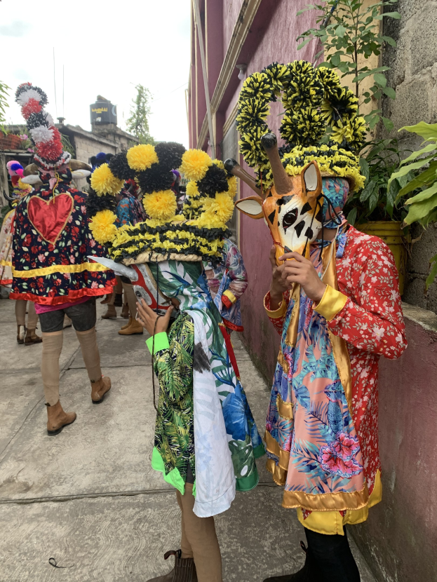 Eine Gruppe von Menschen in bunten Kostümen, einige mit Gegenständen, die zusammen vor einem Gebäude stehen, während eines Faschingsumzugs, mit Bäumen, anderen Gebäuden und einem klaren blauen Himmel im Hintergrund.