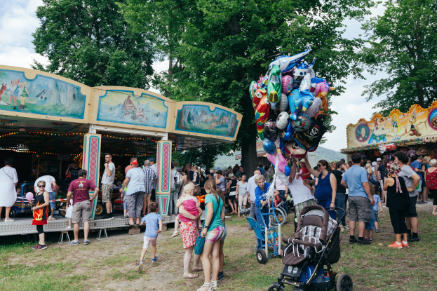 Eine Menschenmenge um einen bunten, beleuchteten Karussellstand an einem Volksfest, einige mit Kinderwagen, mit Bäumen und einem bewölktem Himmel im Hintergrund.