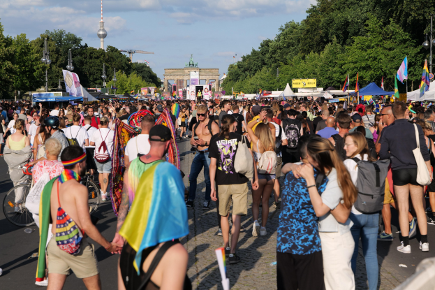 Eine große Menschenmenge marschiert auf einer Straße in Berlin, Deutschland, einige tragen Mützen und tragen Taschen oder Fahnen, mit Zelten, Bannern, Laternenmasten, Bäumen, Gebäuden und einem Turm im Hintergrund unter einem bewölkten Himmel.