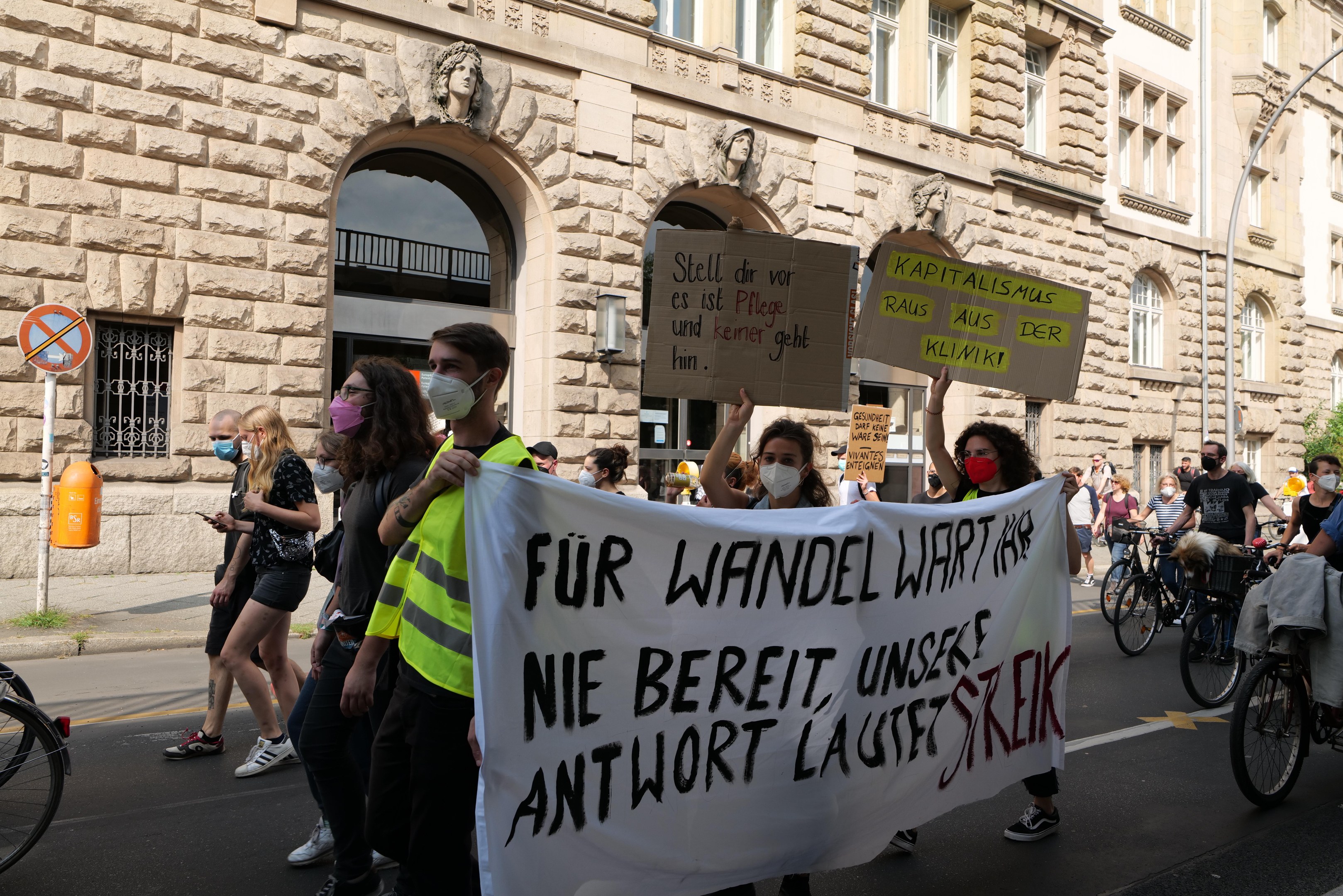 Eine Protestdemonstration auf einer Stadtstraße mit Menschen, die Schilder halten und Fahrräder fahren, an einem Gebäude mit Rundbogenfenstern, Säulen und skulpturalen Details, Bäumen und einer Straßenlaterne vorbei.
