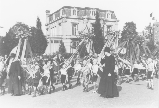 Ein Schwarz-Weiß-Foto einer Gruppe von Menschen, die eine Straße entlanggehen, einige halten Fahnen, tragen Hüte, Bäume, Gebäude und einen sichtbaren Himmel im Hintergrund, mit einem Maibaum als Hinweis auf ein Maifeier.