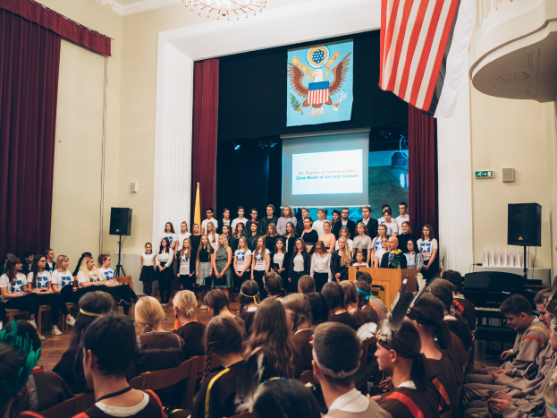 Eine Gruppe von Menschen steht auf einem Podium mit Mikrofon, flankiert von Fahnen und Lautsprechern, und spricht eine sitzende Menge an, während im Hintergrund ein Bildschirm, Vorhänge und ein Kronleuchter zu sehen sind.