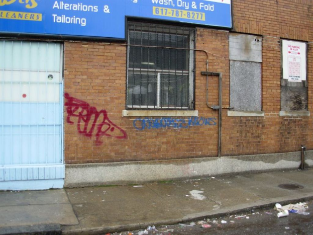 Eine Straßenszene mit einem Gehweg und einer Straße voller Müll, einer Backsteinwand, Fenstern, Rohren, einem Tor und einer Wandtafel oben.