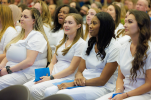 Eine Gruppe von Frauen in weißen Kleidern sitzt in einem Hörsaal, jede hält ein blaues Objekt, mit einem unscharfen Hintergrund, der die Frauen in den Vordergrund stellt, die scheinbar Teil eines Pflegeprogramms sind.