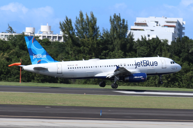 JetBlue Airbus A320-200 beim Start von Miami International Airport mit grünem Gras, hohen Bäumen, Gebäuden im Hintergrund und einem klaren blauen Himmel.