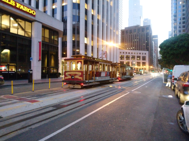 Eine Seilbahn fährt eine Straße in San Francisco entlang, mit Fahrzeugen, einem Fußgänger und einem geparkten Van, vor dem Hintergrund von Gebäuden, Bäumen und Himmel.