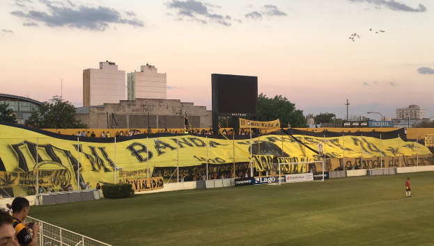 Ein Fußballfeld mit einer Gruppe von Menschen darauf, umgeben von einem Metallzaun, einem Torpfosten, einer Tafel mit der Aufschrift "Boca Juniors vs Real Sociedad", einigen Pflanzen, einem Anzeigebildschirm, einigen Pfosten, der Rinde eines Baumes, einigen Bäumen, einigen Gebäuden und einem bewölkten Himmel.