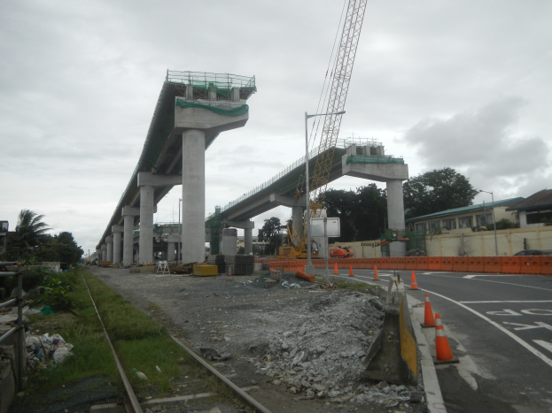 Baustelle mit einer Brücke im Hintergrund, Straße mit Absperrgittern, Bahnschienen, verstreute Steine und Gras, Bäume und Gebäude auf beiden Seiten und ein bewölkter Himmel.