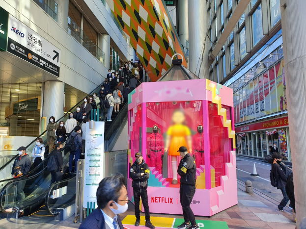 Eine Gruppe von Menschen steht vor einem Gebäude mit Glaswänden, Säulen und Texttafeln, einige tragen Masken, während hinter ihnen eine Rolltreppe mit Menschen nach oben fährt; ein pinker Kasten in der Mitte zeigt Schaufensterpuppen und trägt die Aufschrift "Netflix's neuer Netflix-Stand in Tokyo."