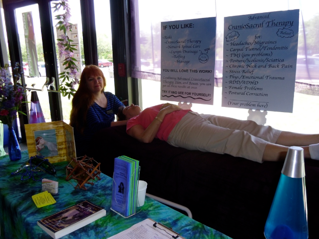 Eine Frau liegt auf einem Therapietisch, während eine andere Frau eine Therapiestunde durchführt, mit Postern, die die Therapie beschreiben, und einem Tisch mit Gegenständen und Büchern daneben.