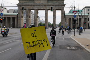 Eine Gruppe von Radfahrern in Helmen fährt auf einer Straße am Brandenburger Tor in Berlin, Deutschland vorbei, wobei einer ein gelbes Schild hält, unter einem klaren blauen Himmel mit Gebäuden und Bäumen im Hintergrund.