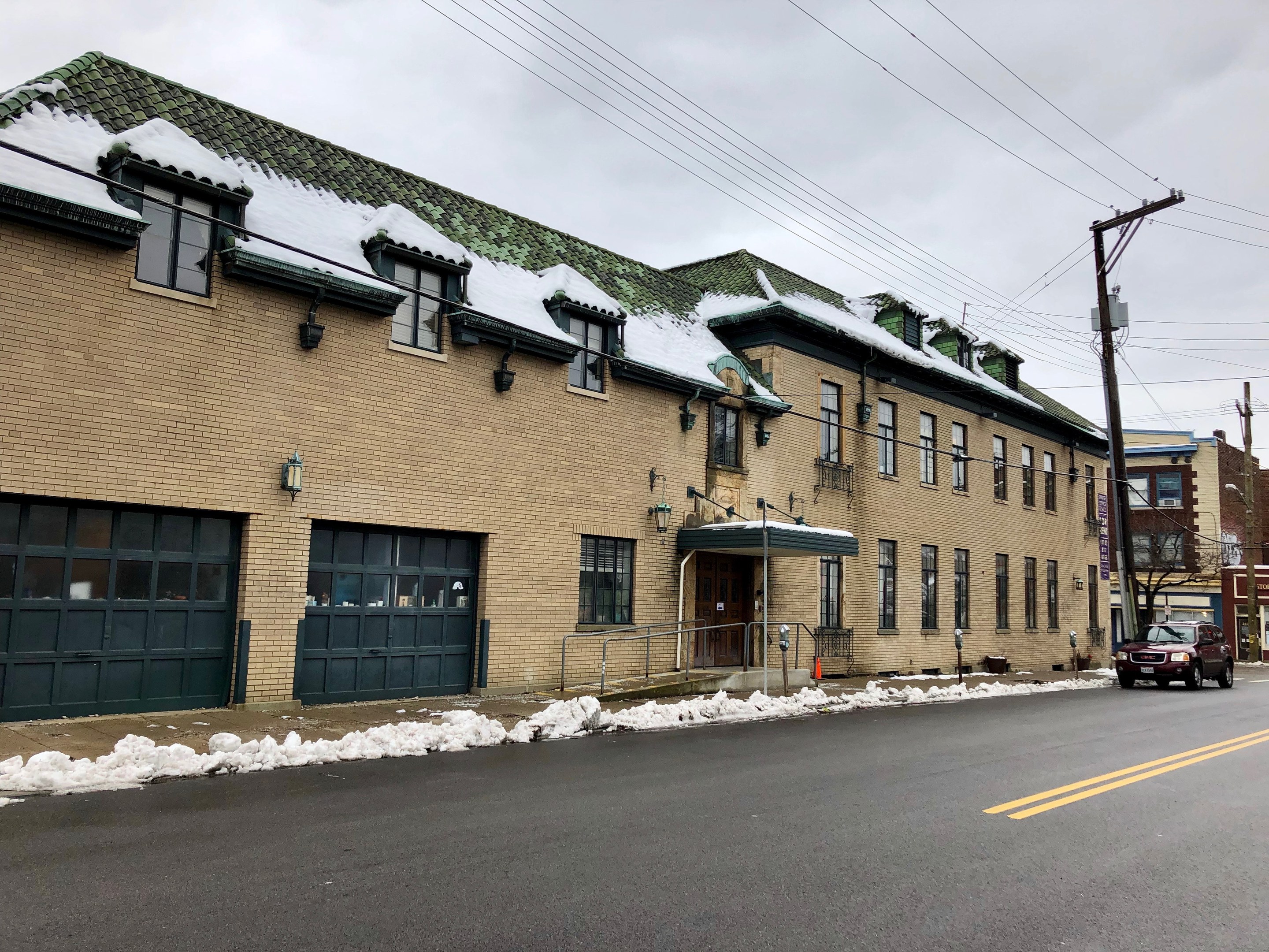 Großes Backsteingebäude mit grüner Dachfläche an einer Straßenkreuzung, umgeben von Fahrzeugen, Schnee, Strommasten, Bäumen und einer bewölkten Himmel, identifiziert als die ehemalige Feuerwache St. Louis.