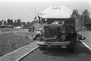 Ein Schwarz-Weiß-Foto eines schwer beschädigten Autos am Straßenrand mit eingedellter Motorhaube und zersplitterten Fenstern, anderen Fahrzeugen, Laternenpfählen, Bäumen und einem Haus im Hintergrund.