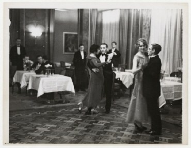 Schwarzes und weißes Foto eines Paares, das in einem Ballsaal tanzt, mit Zuschauern an Tischen sitzend, Vorhängen im Hintergrund und einem Rahmen an der Wand.