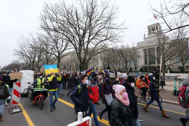 Eine große Gruppe von Menschen nimmt an einer Protestdemo auf den Straßen von Washington, D.C. teil, hält Schilder und Banner hoch, fährt Fahrräder und trägt Schilder mit Stöcken und Bäumen unter einem klaren blauen Himmel.