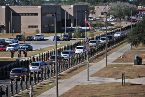 Eine große Gruppe von Menschen in militärischer Kleidung mit Gewehren vor einem Gebäude während einer Parade, mit Fahrzeugen, Laternenmasten, Flaggen, Bäumen und Gras im Vordergrund und Gebäuden unter einem klaren blauen Himmel im Hintergrund.