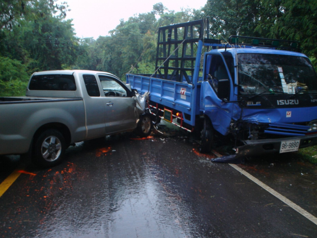 Ein schwer beschädigtes LKW-Wrack mit eingedrüteter Front und verbeulter Karosserie liegt am Straßenrand, umgeben von Bäumen unter einem klaren blauen Himmel.