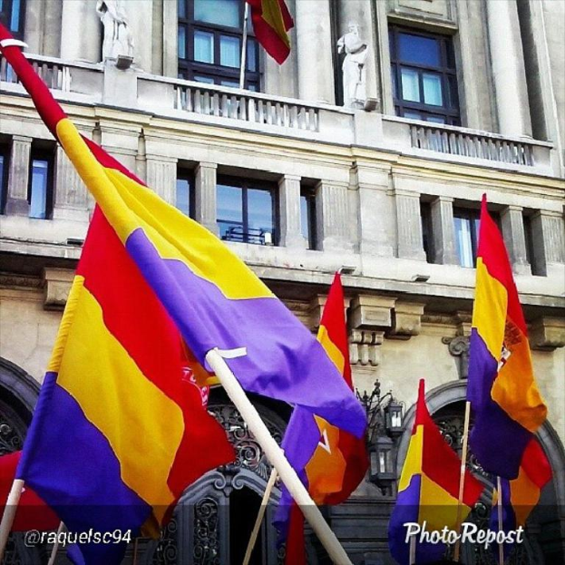 Ein mehrstöckiges Gebäude mit zahlreichen Fenstern und Türen, mit mehreren Masten davor, an denen Flaggen in Rot, Gelb und Lila wehen.