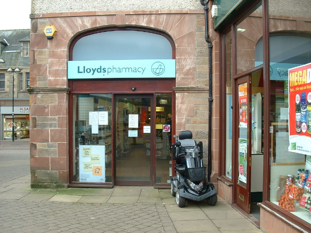 Eine Apotheke mit einem vor ihr geparkten Fahrzeug und einem Gebäude links im Bild.