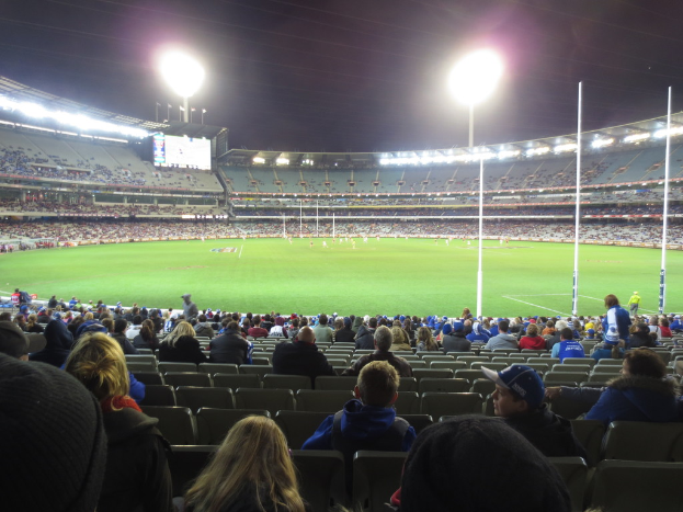 Ein Stadion mit vielen Menschen auf Stühlen auf einem Rasen, mit Leuchten im Hintergrund und auf der rechten Seite und einem Bildschirm in der Ferne.