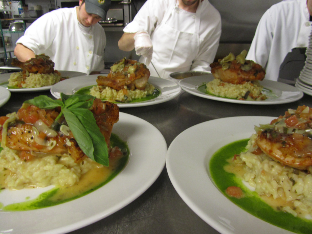Drei Köche in einer Küche mit Mützen und Handschuhen bereiten Teller mit Speisen zu, mit Regalen im Hintergrund.