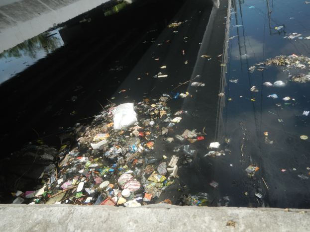 Ein mit Müll gefüllter Fluss neben einer Brücke, mit einer Wand auf der linken Seite und einer Straße unten.