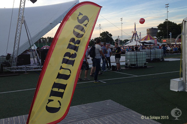 Eine lebhafte Karnevalsszene mit mehreren Zelten, einem zentralen Mast und Menschen unter den Zelten, umgeben von Bäumen, Strommästen, Straßenbahnen auf der Straße und einem eisernen Ständchen auf der linken Seite, unter einem klaren Himmel.