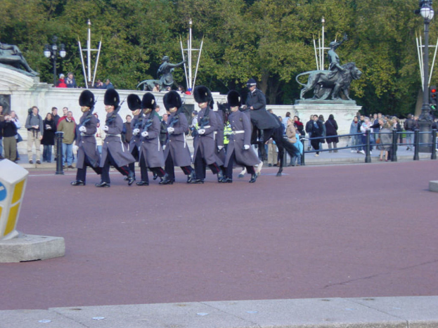 Eine Gruppe von Soldaten in grauen Uniformen marschiert mit Gewehren, ein Mann zu Pferd neben ihnen, Zuschauer schauen zu, Statuen an einer Wand und Bäume im Hintergrund.