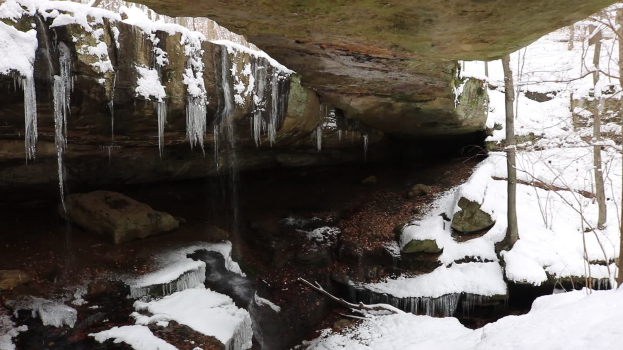 Ein kleiner Wasserfall ergießt sich an einem schneebedeckten, steinigen Hang in einem wöldernen Gebiet, mit Eiszapfen, die von den Felsen hängen und von Schnee bedeckten Bäumen umgeben sind.