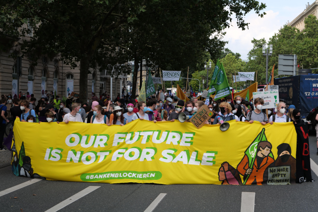 Eine große Gruppe von Menschen, die eine Straße entlanggehen und ein Transparent mit der Aufschrift "Unsere Zukunft ist nicht zu verkaufen" tragen, mit Bäumen, Laternenmasten und Gebäuden im Hintergrund unter einem bewölkten Himmel.