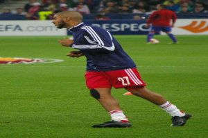 Ein Mann in einem blauen Hemd und roten Shorts läuft auf einem Fußballfeld, im Hintergrund sitzt eine Gruppe von Menschen und es sind einige Werbetafeln sichtbar.