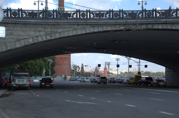 Eine Brücke überspannt eine vielbefahrene Straße, unter der Fahrzeuge hindurchfahren, während Fußgänger auf dem Gehweg links stehen, mit städtischen Elementen wie Verkehrsampeln, Bäumen, Gebäuden und Laternen im Hintergrund.