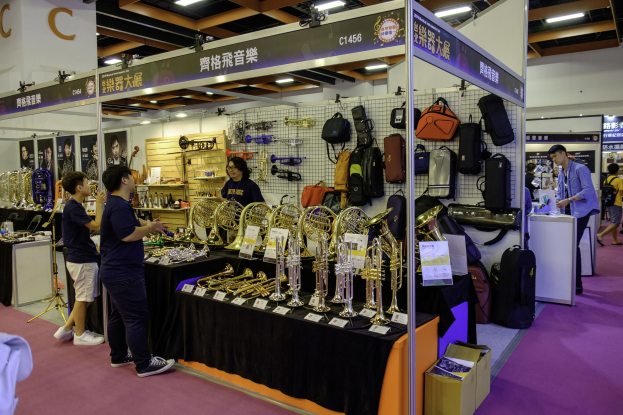 Gruppe von Menschen um einen Tisch mit Musikinstrumenten, Taschen und Tafeln mit Text bei dem Hong Kong International Music Festival, mit Deckenbeleuchtung und einem Karton auf dem Boden.