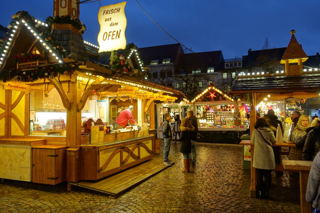 Ein belebter Weihnachtsmarkt auf einer Kopfsteinpflasterstraße bei Nacht mit Menschen um beleuchtete Stände, Gebäuden, Bäumen und einem klaren Himmel im Hintergrund.